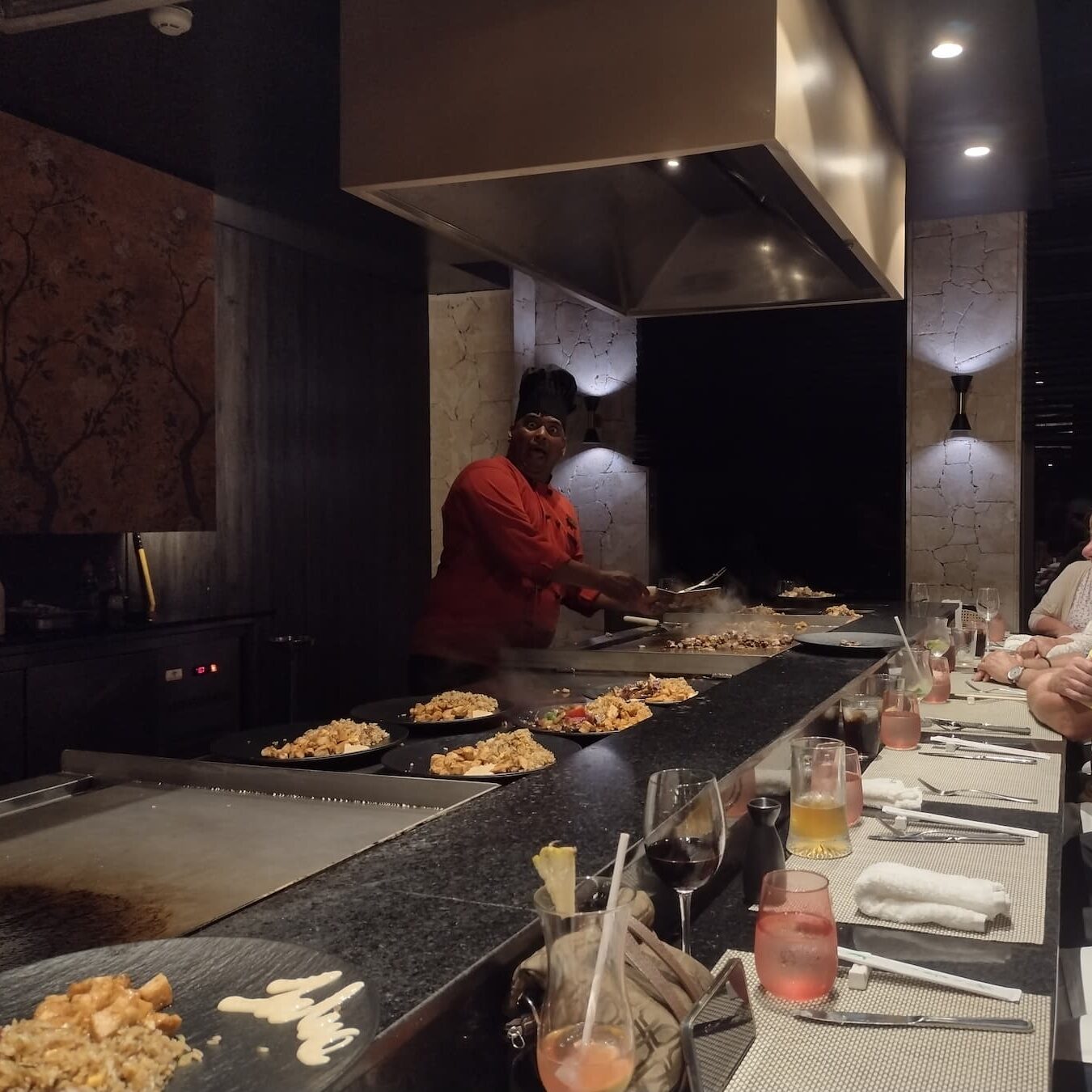 Chef preparing food on a sizzling hibachi grill at Himitsu restaurant in Dreams Flora, with flames and flying shrimp entertaining guests.